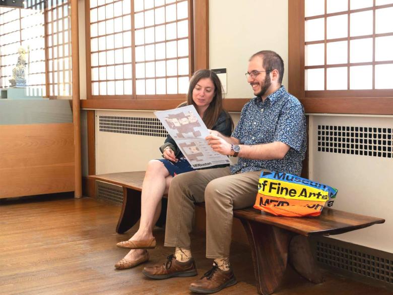 Two people sit on an orange and deep brown wooden bench in a brightly lit hallway.
