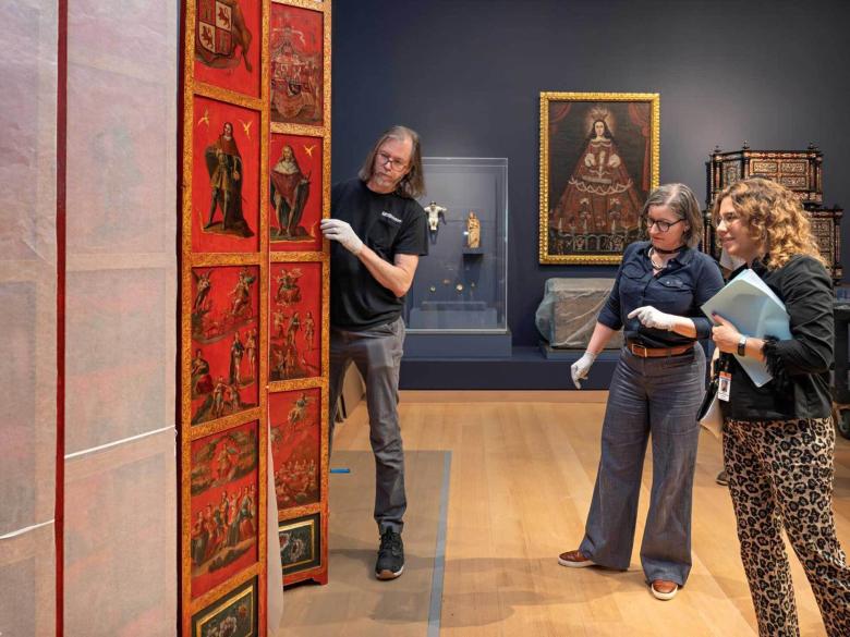 Three people stand in a dark room and look at a bright orange folding screen.