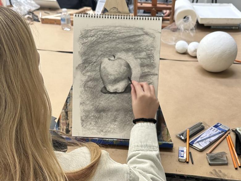 Teen student draws an apple in charcoal on table top easel in classroom (SAC)