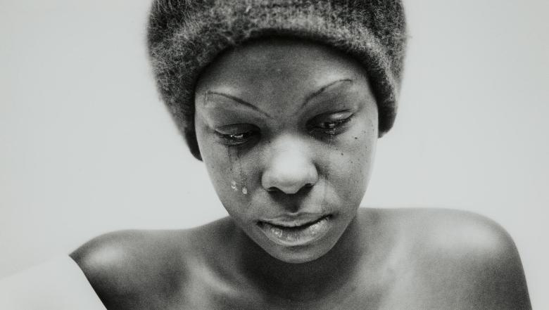 A woman shown from the shoulders up looks down as tears fall from her eyes.