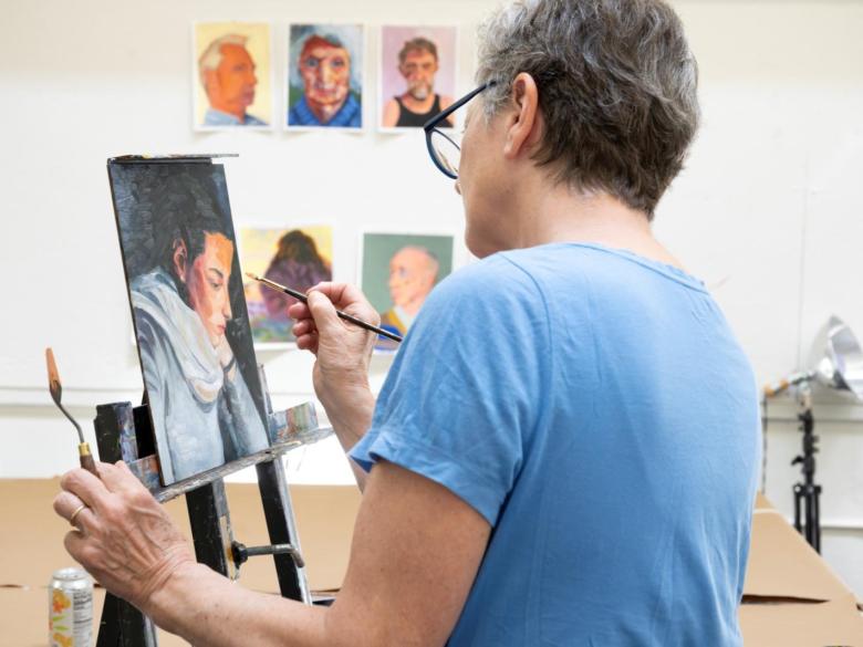 adult student paints a portrait on easel in studio classroom with paintings in background (SAC)