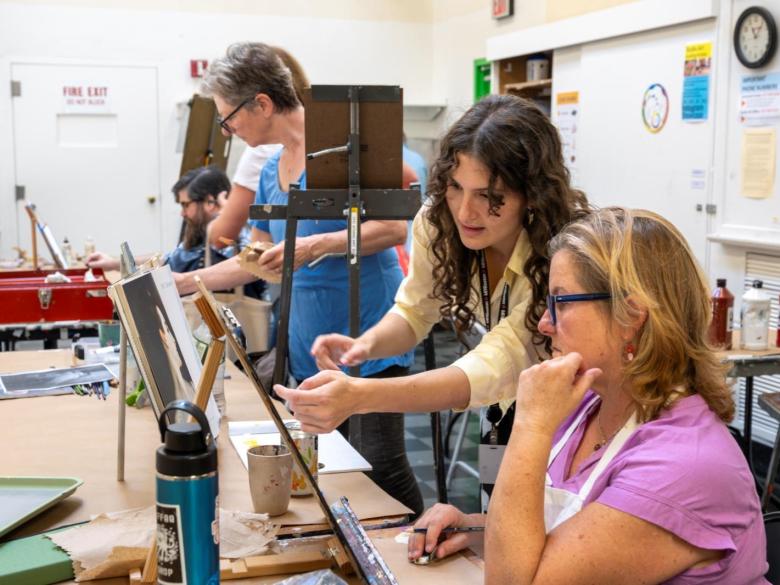 Studio Art Instructor points at table top easel with students painting in classroom (SAC)