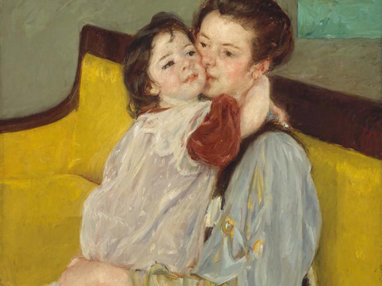 A young child tenderly clings to a woman sitting on a yellow sofa.